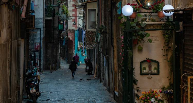 Smalle geplaveide steeg in Napels omzoomd met hoge verweerde gebouwen, scooters en hangende lampen
