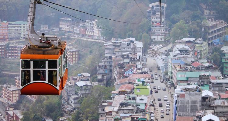 Eine orange Seilbahn fährt über eine Stadtlandschaft am Hang mit Verkehr und Gebäuden darunter.