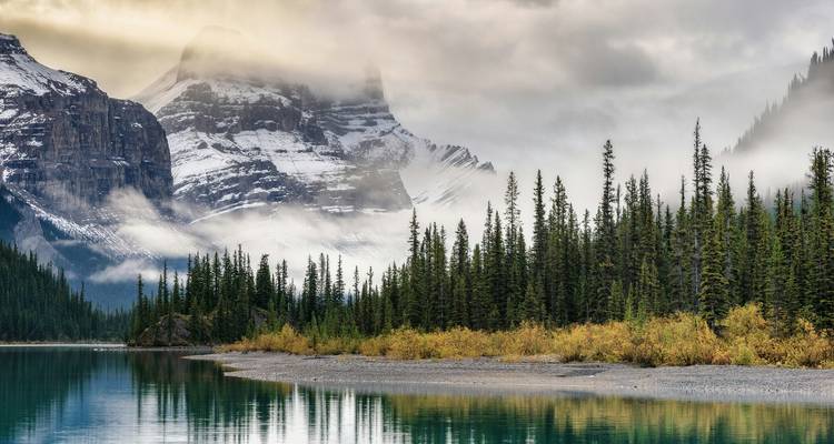 Snow-capped Rocky Mountains reflected in a calm turquoise lake bordered by evergreen forest.