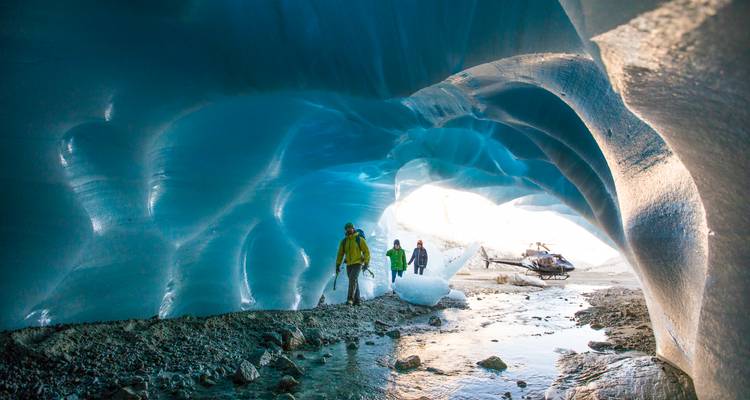 Adventurers exploring a glowing blue ice cave with a helicopter parked outside the glacier.