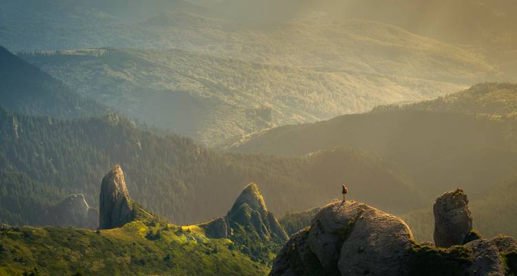 Lone figure standing atop rugged rock spires overlooking misty forested valleys at golden hour.