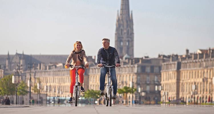 Hombre y mujer en bicicleta atravesando una gran plaza en Burdeos con torre gótica de fondo.