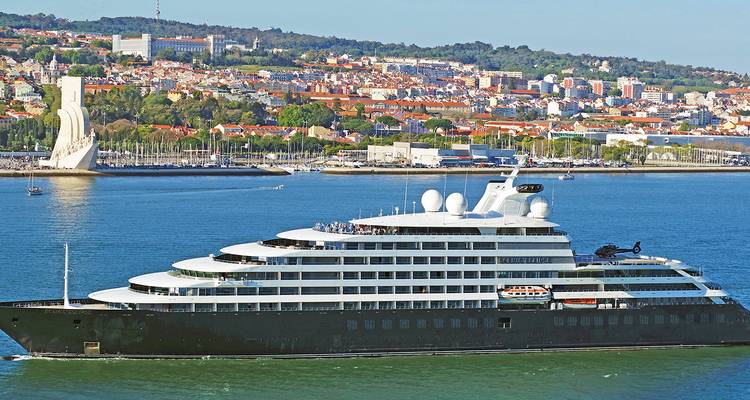 Gran crucero moderno navegando junto a una ciudad costera bajo cielos azules despejados.