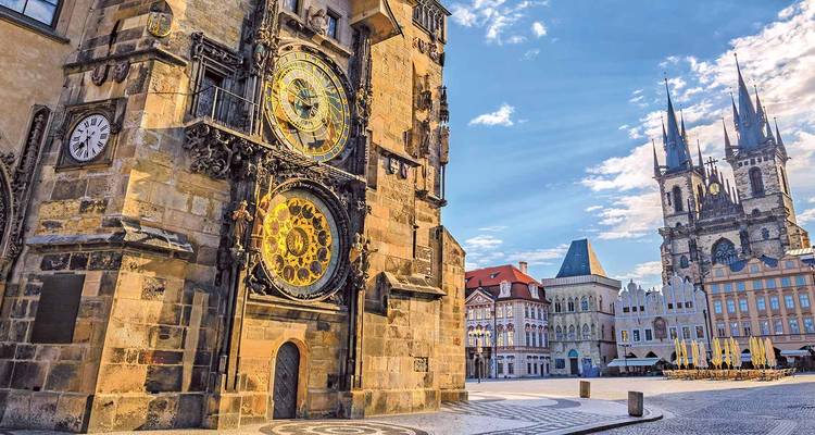 Primer plano de la torre del Reloj Astronómico de Praga con las torres de la Iglesia de Týn bajo un cielo azul.