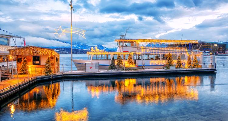 Bateau de croisière illuminé de façon festive et cabane en bois se reflétant sur les eaux calmes du lac de Lucerne au crépuscule avec des sommets enneigés au loin.
