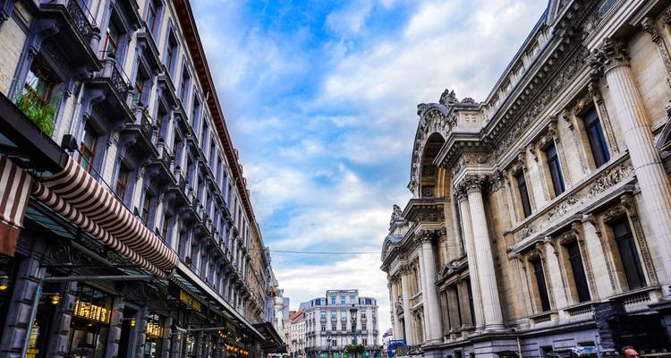 Grandes fachadas clásicas que bordean una concurrida calle central en Bruselas bajo un cielo parcialmente nublado.