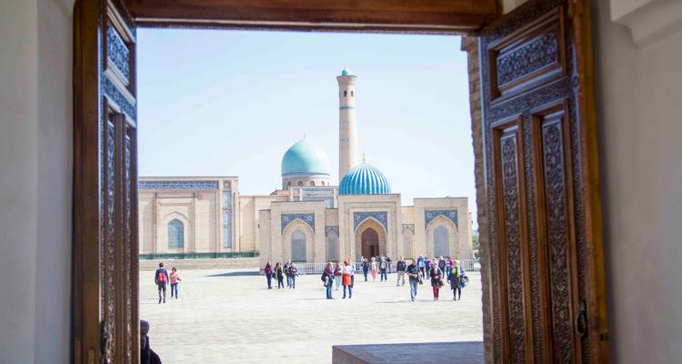 Foule de visiteurs entrant dans le complexe Hazrati Imam vue à travers une porte en bois sculptée de manière ornée.