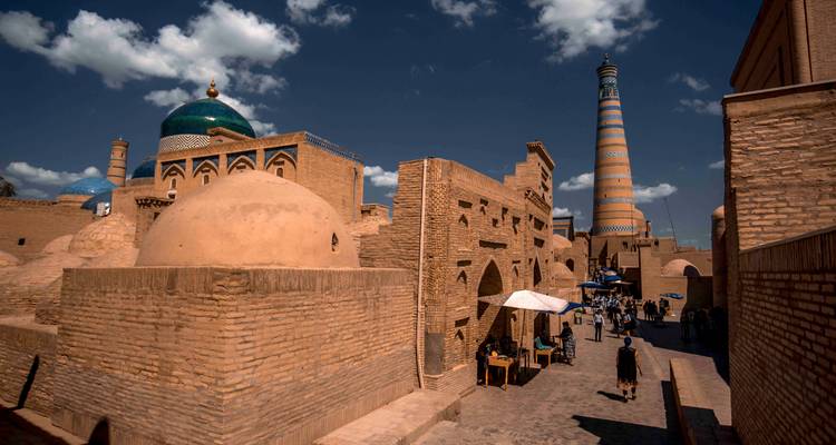 Rue animée en adobe à Khiva avec des parasols de marché, des dômes et un minaret rayé sous des nuages spectaculaires.