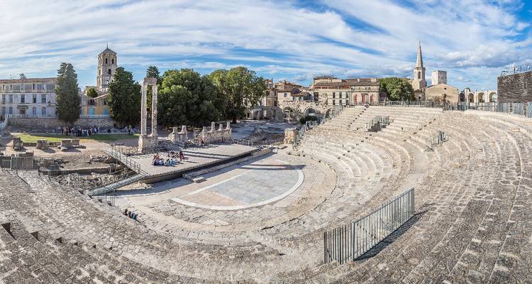 Panoramablick auf das antike römische Amphitheater und die umliegenden historischen Gebäude in Arles unter einem strahlend blauen Himmel mit streifigen Wolken.
