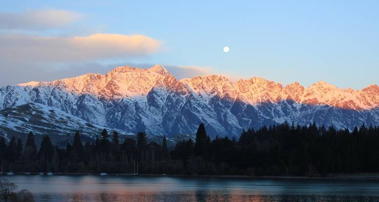 Besneeuwde bergketen die roze gloeit in het vroege ochtendlicht boven een kalm meer en bos in Queenstown.