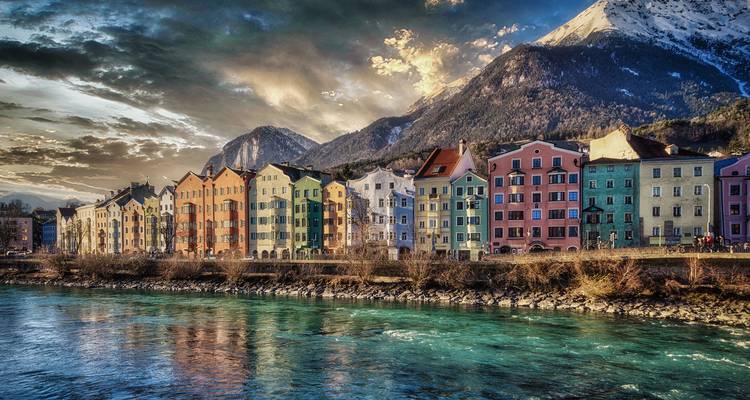 Colorful riverside houses in Innsbruck sit beneath snow-dusted Alpine peaks at sunset.