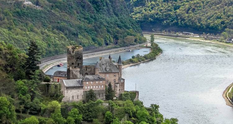 A medieval castle perches above the Rhine River surrounded by lush green hills.