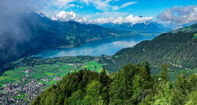 A bright blue alpine lake is surrounded by mountains, forests, and patchwork farmland.