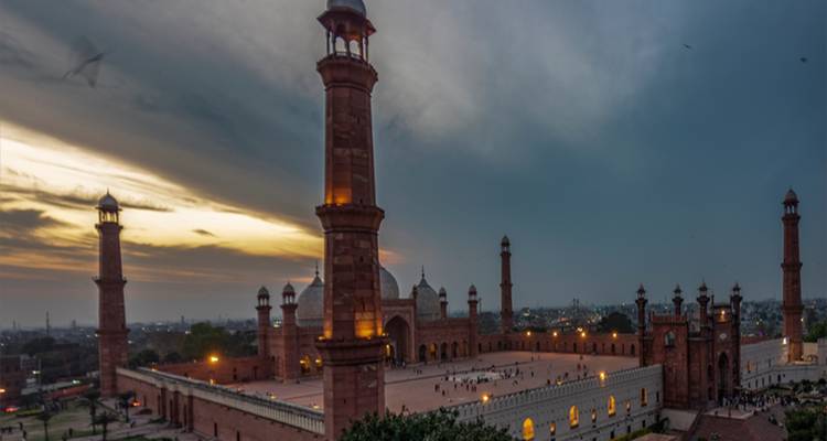 Große rote Sandsteinmoschee mit mehreren Minaretten unter dramatischem Abendhimmel.