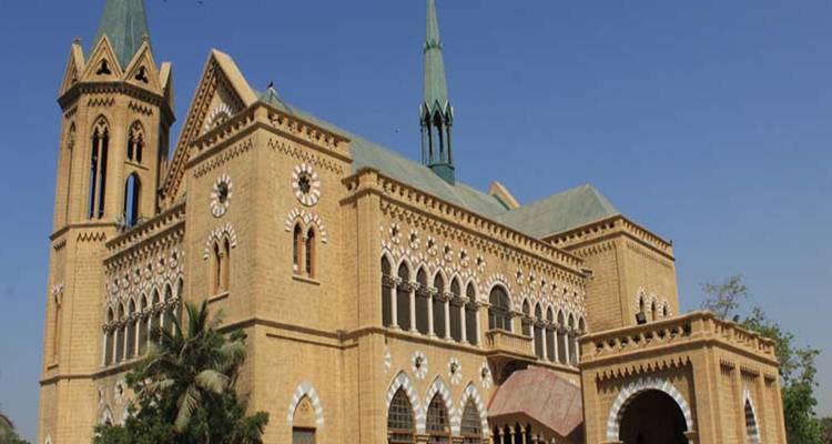 Gotische gelbe Sandsteinhalle mit Bogenfenstern und hohem Turm vor klarem Himmel.