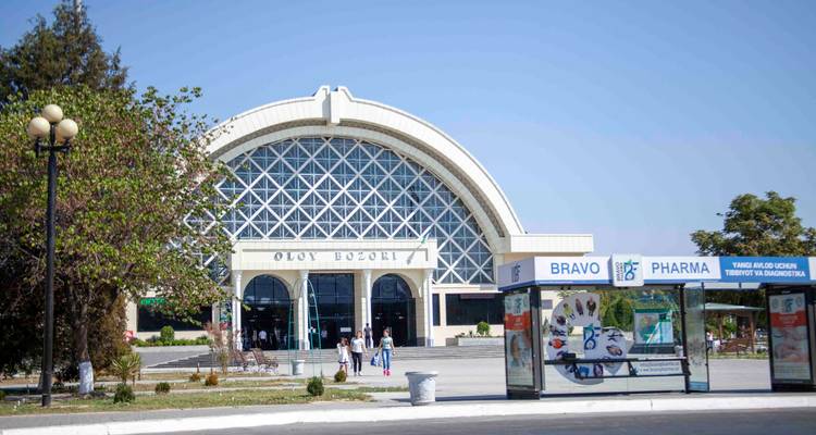 Bâtiment de bazar moderne à arcades 'Oloy Bozori' avec des gens qui marchent et un arrêt de bus devant.