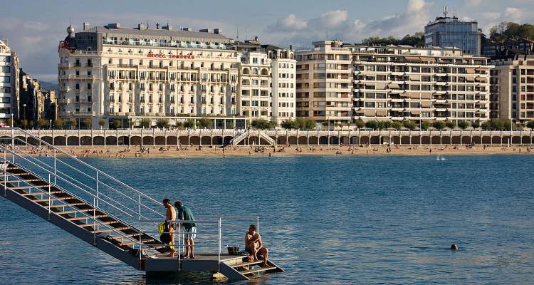 Escena de playa con nadadores en una escalera metálica que lleva al mar y grandes edificios junto al mar de fondo.