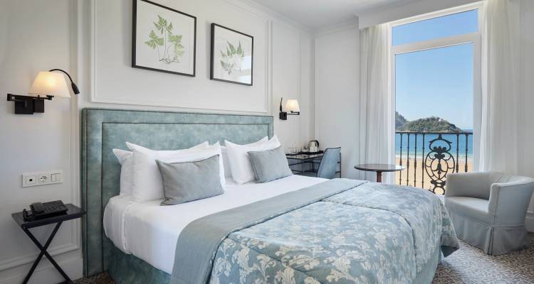 Habitación de hotel elegante con decoración en tonos pastel, balcón y vista al mar.
