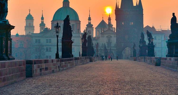 Vroege ochtendaanblik langs de Karelsbrug in Praag met standbeelden als silhouetten tegen een oranje zonsopgang en verre koepels.
