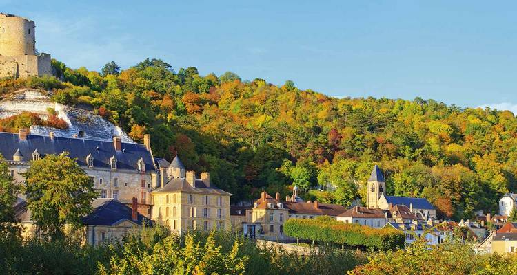Mittelalterliche Burgruinen auf einem Hügel mit Blick auf eine farbenfrohe bewaldete Stadt unter strahlend blauem Himmel.