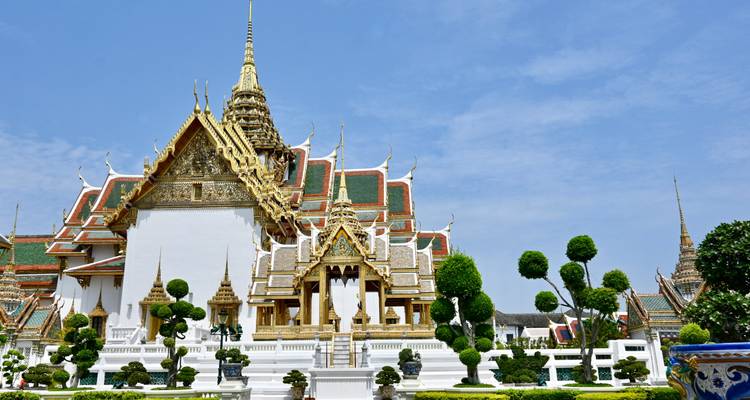 Complexe du Grand Palais avec des toits thaïlandais ornés, des flèches dorées et des topiaires parfaitement entretenues sous un ciel bleu clair.