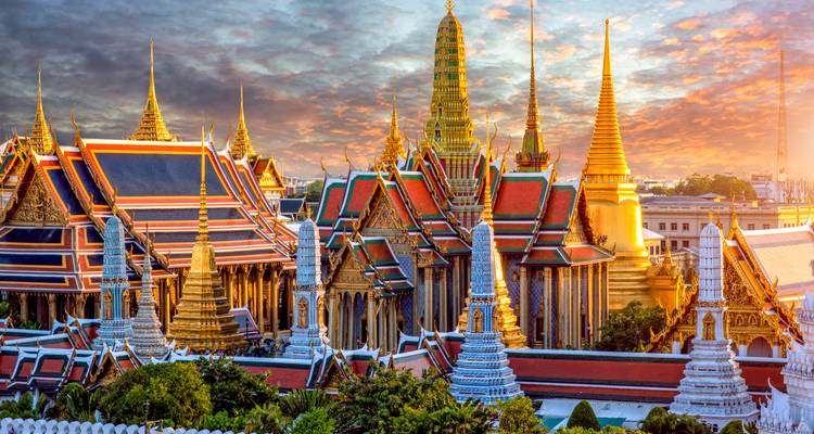 Bunte Ziegeldächer und vergoldete Turmspitzen des Grand Palace in Bangkok unter einem dramatischen Sonnenaufgangshimmel.