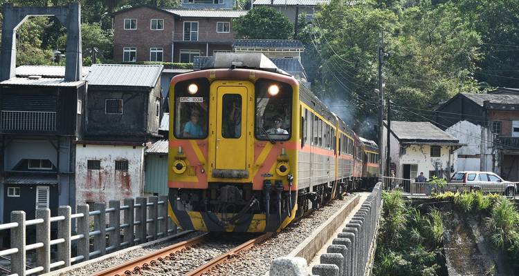 Gele lokale trein die een smalle brug oversteekt te midden van landelijk Taiwanees dorpslandschap.