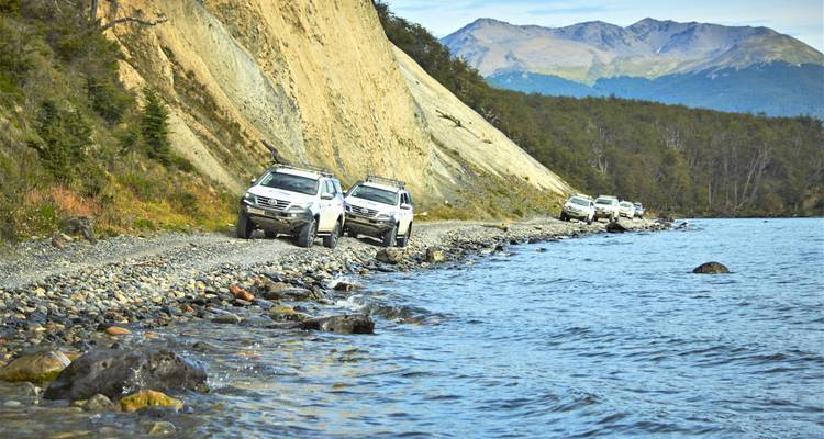 Konvooi van terreinvoertuigen die langs rotsachtige oever van meer rijden aan de voet van beboste kliffen.
