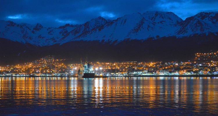 Avondpanorama van de stadsverlichting van Ushuaia weerkaatst op kalm water met besneeuwde pieken als silhouet.