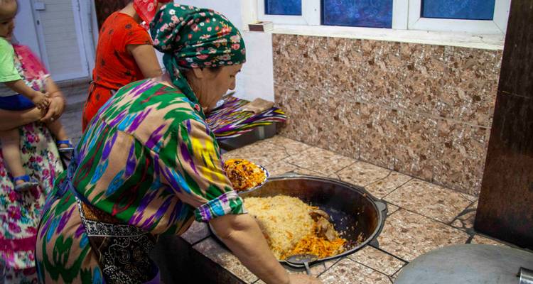 Femme en robe colorée servant du riz d'un grand kazan pendant que la famille regarde.