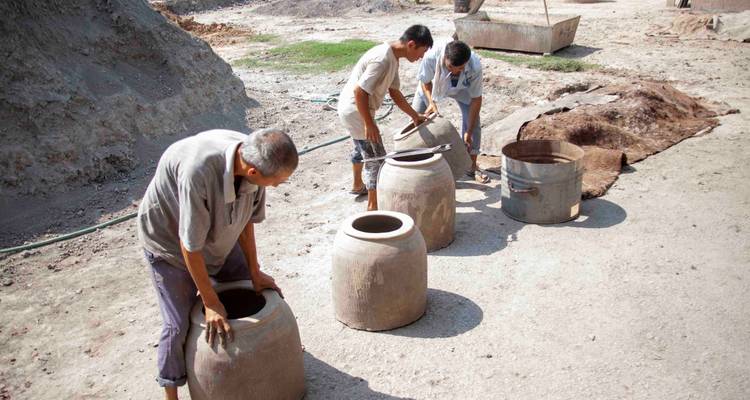 Trois hommes penchés au-dessus de grandes jarres en argile lors d'une cuisson traditionnelle de poterie en plein air.