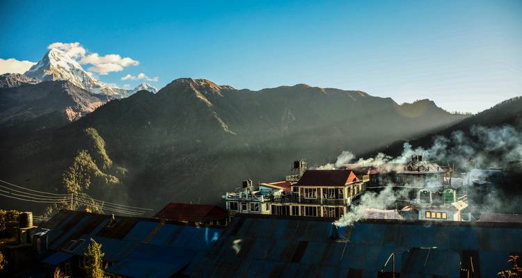 Mountain village rooftops with chimneys smoking beneath the dramatic Annapurna range at golden sunrise.