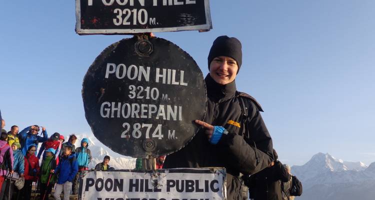 Hiker smiles beside the Poon Hill altitude sign with distant snow peaks and fellow trekkers behind.