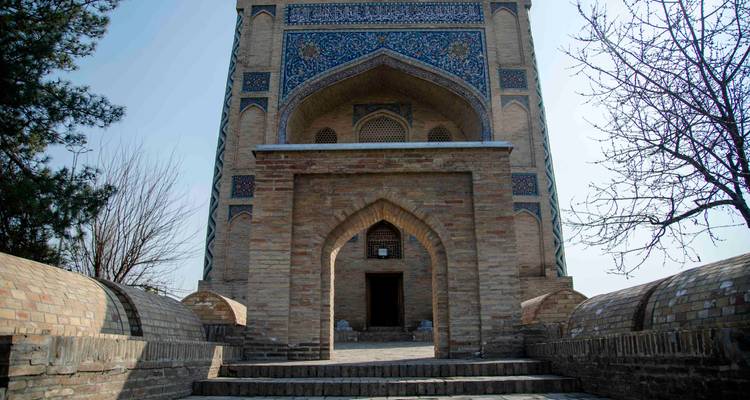 Portail en brique orné de mosaïques bleues vu depuis une allée pavée ombragée et des marches.