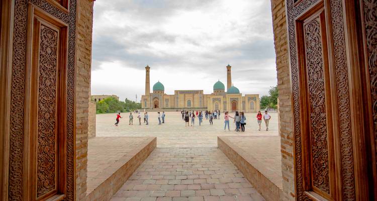 Scène de cour de Hazrati Imam avec des touristes vue à travers des portes en bois richement sculptées sous un ciel couvert.