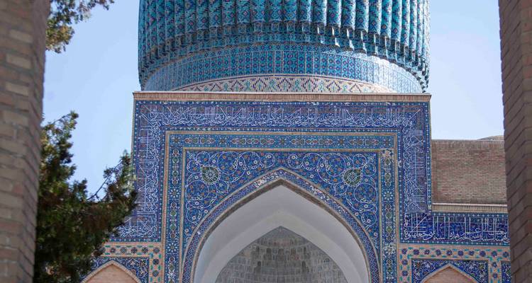 Gros plan du portail richement carrelé et du dôme turquoise à nervures qui dépasse entre les arbres.