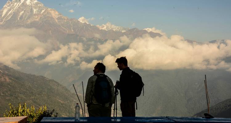 Two hikers with backpacks stand on a ridge looking over cloud-filled Himalayan valleys.