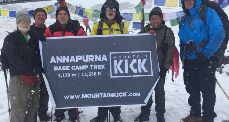 Trekking team holding a banner reading 'Annapurna Base Camp Trek' amid falling snow.