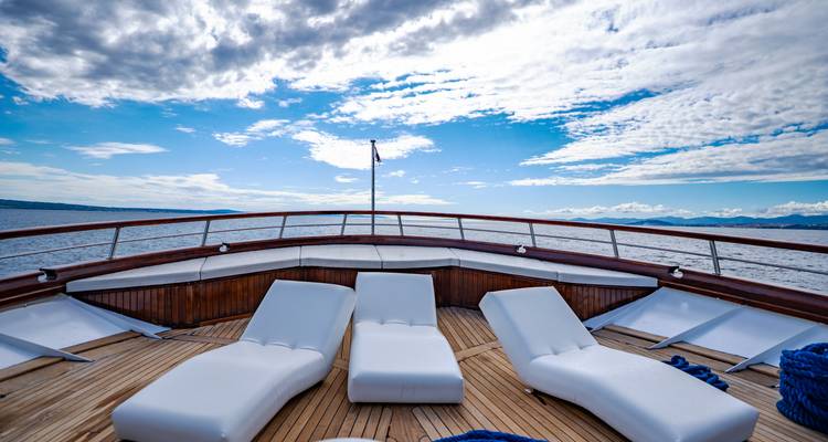 Bow deck of a yacht with white loungers facing out to sea under a patterned sky.