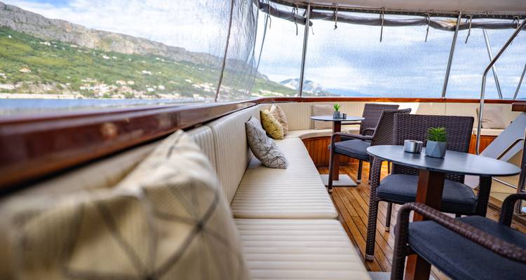 Side bench and small tables on a yacht with coastal scenery beyond.