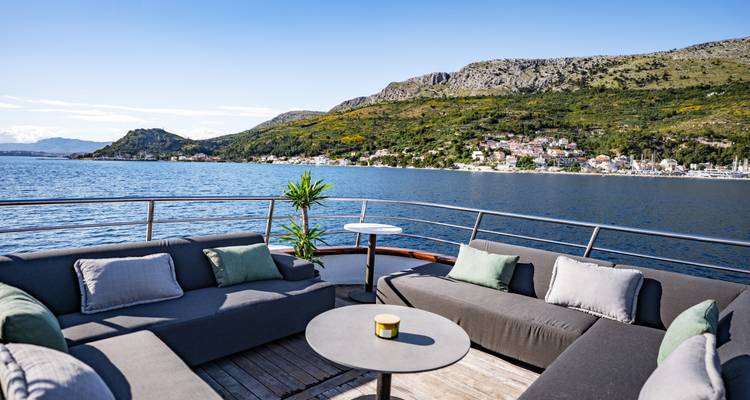 Outdoor lounge seating on yacht with coastal town and hills in the distance.