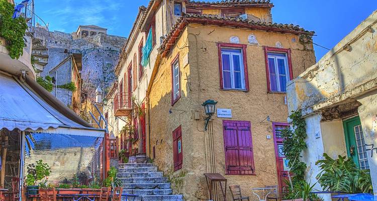 Calle angosta y colorida en el barrio Plaka de Atenas con mesas de café y casas antiguas.