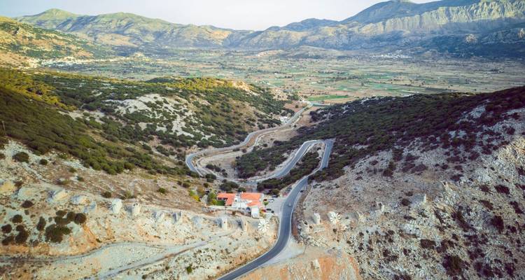 Un sinuoso camino de montaña serpentea a través de colinas rocosas áridas hacia un valle fértil en Creta.