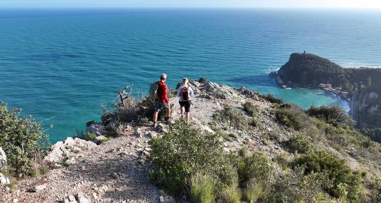 Two hikers stand on rocky promontory overlooking wide expanse of blue Mediterranean Sea coastline