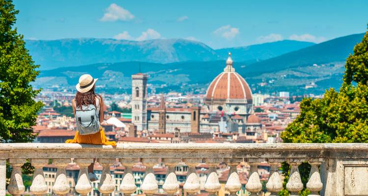 Voyageuse avec chapeau et sac à dos assise sur la rambarde d'une terrasse, admirant l'horizon de Florence dominé par le Duomo par une journée ensoleillée.
