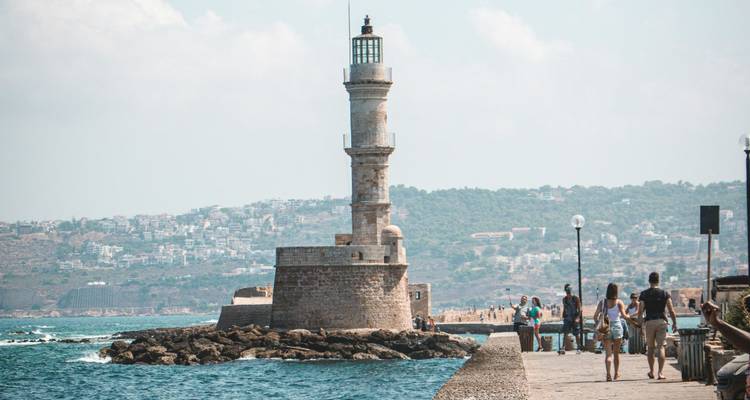Faro histórico de estilo egipcio que se alza sobre el rompeolas del puerto con visitantes paseando cerca.