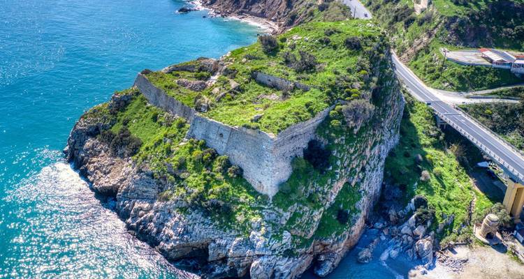 Fortaleza en ruinas en la cima de una colina cubierta de vegetación con vista a una costa rocosa y un mar azul profundo.