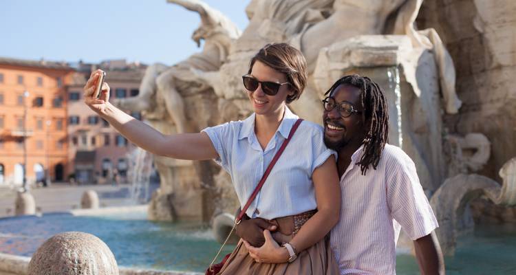Couple souriant prenant un selfie devant une fontaine baroque à Rome par un après-midi ensoleillé.