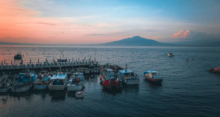 Fishing boats bob on calm water at sunset with Mount Vesuvius silhouetted in the distance.