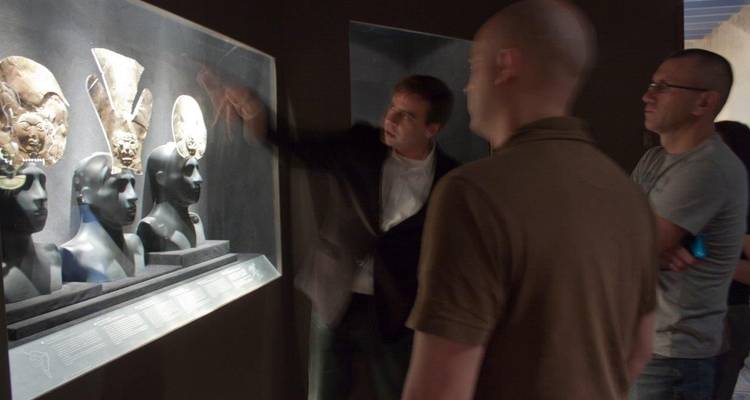 Guide de musée pointant des artefacts en or antiques dans une exposition faiblement éclairée tandis que deux visiteurs observent